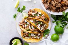 three shredded pork tacos on a plate with limes and cilantro next to it