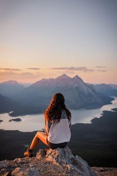 a woman sitting on top of a rock next to a lake at sunset with mountains in the background