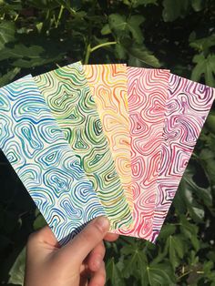 hand holding four different colored papers in front of green bushes and shrubbery behind them