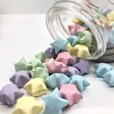 small pastel origami stars are scattered around a glass jar on a white surface