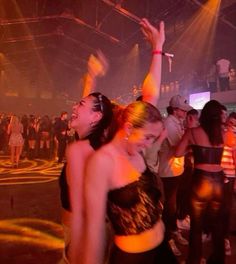 two young women dancing at a party with other people in the background and lights on