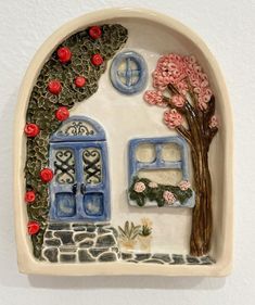 a ceramic wall hanging with flowers and a tree in the window, on a white wall