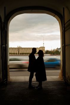 two people standing under an archway with cars passing by