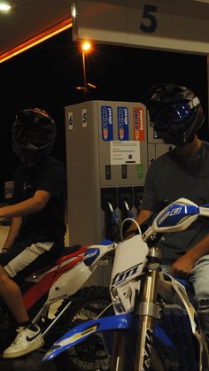 two people are sitting on motorcycles at a gas station, one is wearing a helmet