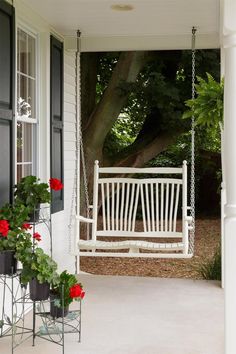 a white porch swing sitting next to a tree and potted plants on the front porch