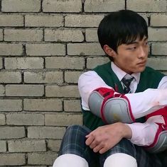 a young man sitting against a brick wall wearing a green vest and white socks with his arms crossed