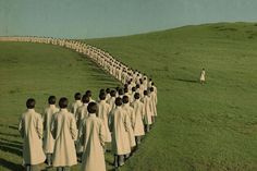 a long line of people in white coats walking across a grass covered field next to each other