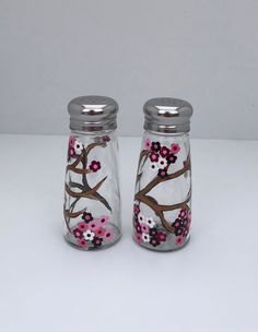 two glass jars with pink flowers painted on the side and silver lids are sitting on a white surface