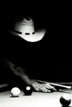 a man in a cowboy hat leaning over a pool table with cues and balls