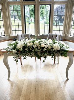 a table with white flowers and greenery on it in front of two large windows