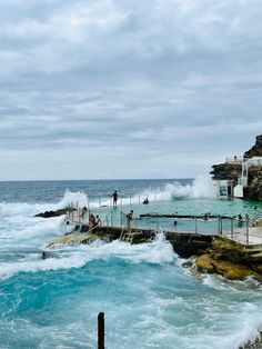 people are standing on the edge of an ocean pool as waves crash in the background