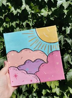 a hand holding up a small painting in front of some bushes and plants with the sun above it