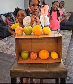 two girls standing behind a cardboard box with apples and oranges in it while another girl looks on