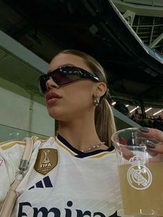 a woman wearing sunglasses and holding a drink in her hand at a sporting event with people watching from the bleachers