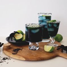 two glasses filled with black liquid and limes on a wooden cutting board next to other drinks