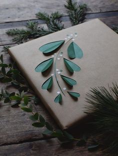 a gift wrapped in brown paper with green leaves and pearls on the side, surrounded by greenery