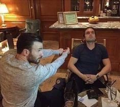 two men sitting at a table with wine glasses in front of them and one man holding his hand up to his face