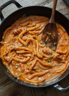 a skillet filled with pasta and sauce on top of a wooden table next to a spoon