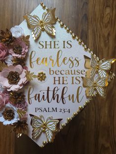 a white graduation cap with gold lettering and pink flowers on the side that says, she is fearless because he is faithful