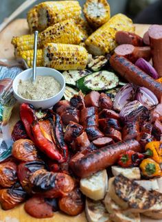 an assortment of grilled meats and vegetables on a wooden platter with a bowl of dip