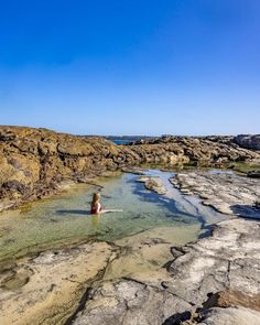 a person standing in the middle of some water on rocks and sand with blue skies above