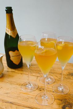 three glasses filled with yellow liquid next to a bottle of champagne on a wooden table