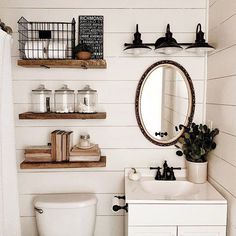 a white toilet sitting next to a sink in a bathroom under a wooden shelf filled with bottles