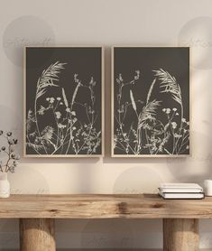 two framed art prints on a wall above a wooden table with books and vases