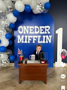 a boy sitting at a desk in front of a blue and white backdrop with balloons