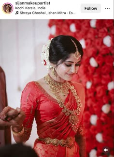 a woman in a red sari and gold jewelry looks down at her hand while standing next to a flower wall