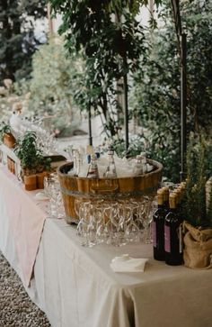 an outdoor table with wine glasses and bottles on it, surrounded by greenery in the background