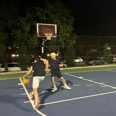 two young men playing basketball on an outdoor court at night with the lights turned on