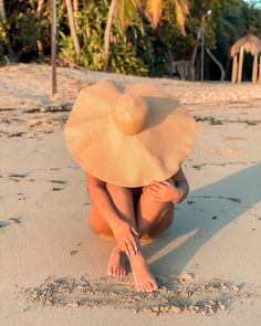 a woman kneeling down on top of a sandy beach wearing a hat and holding her hands behind her head