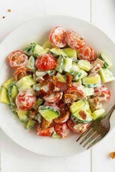 a white plate topped with cucumber, tomatoes and bacon salad next to a fork