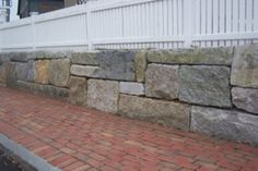 a white picket fence next to a stone wall and brick sidewalk in front of a house