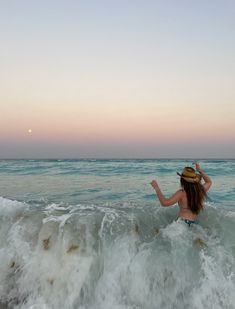 a woman standing in the ocean with her hat on top of her head and arms outstretched