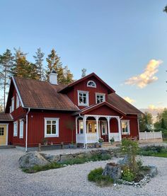 a red house with white trim sits on gravel in front of some trees and rocks