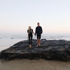 a man and woman holding hands while standing on rocks at the beach