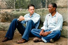 two men sitting next to each other in front of a stone wall with an advertisement on it