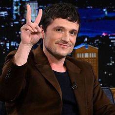 a male in a brown jacket and black shirt is making the peace sign with his hand