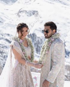 a man and woman standing next to each other in front of snow covered mountains with leis around their necks