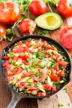 a skillet filled with vegetables sitting on top of a wooden cutting board next to tomatoes and avocado