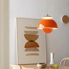 an orange lamp hanging over a wooden table next to a white vase and painting on the wall