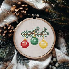 a cross stitch christmas ornament hanging from a pine tree with cones and ornaments