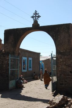 a woman walking down a dirt road under an arch with a cross on it's side