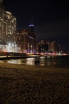 two people walking on the beach at night in front of some tall buildings and lights