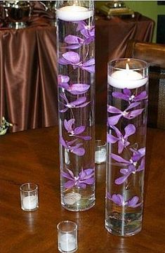 two tall vases filled with water and purple flowers on top of a wooden table