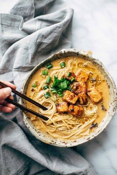 someone is holding chopsticks over a bowl of noodles with tofu and scallions