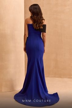 the back of a woman in a long blue dress standing against a wall with her hands on her hips