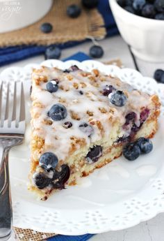 a slice of blueberry coffee cake on a white plate with a fork and bowl of blueberries in the background